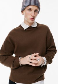 Brązowy sweter z dzianiny z okrągłym dekoltem, nałożony na białą koszulę w paski. Modelka nosi brązową i niebieską czapkę w paski oraz kilka pierścionków.