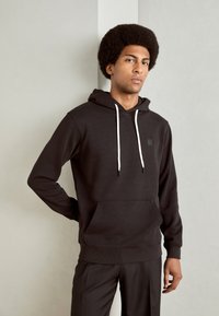 Un jeune homme avec une coiffure afro porte un sweat à capuche noir avec des cordons blancs et un pantalon noir, se tenant devant un mur de couleur claire.