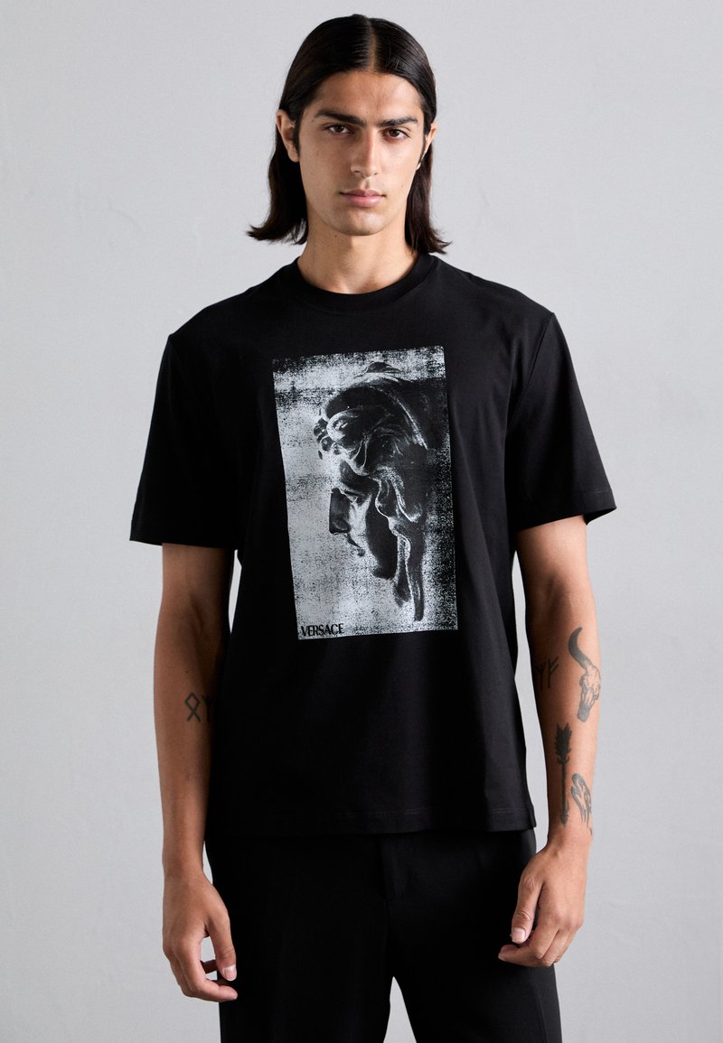 T-shirt en coton noir avec un imprimé graphique en gris et blanc représentant le profil d'un visage, avec le logo "Versace" en bas. Manches courtes.