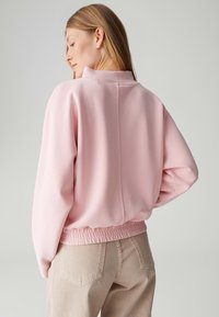 Roze oversized sweatshirt met een hoge hals, elastische zoom en een gladde textuur. Draagt lichtbeige jeans. Naden met detaillering aan de achterkant.