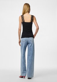 Svart tanktopp med blonderemmer, kombinert med vide, lyseblå jeans. Bakview viser detaljer på skuldre og øvre rygg. Røde hæler fullfører antrekket.