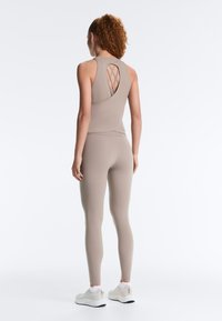 Vrouw met krullend haar in taupekleurige trainingsset met gekruiste rugdetails, witte sneakers aan, staand met de rug naar voren op een witte achtergrond.