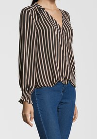 Černá a béžová pruhovaná zavinovací blůza s dlouhými rukávy a nařasenými manžetami, se zvýrazněným hlubokým výstřihem do V a volným střihem. V kombinaci s modrými džíny.