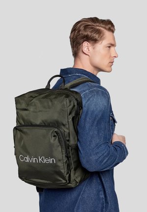 Zaino in nylon verde oliva con più scomparti con cerniera e una tasca frontale, con il logo bianco "Calvin Klein", indossato sulla spalla.