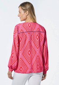 Losse blouse met lange mouwen, voorzien van een geometrisch ruitpatroon in tinten roze en rood, met een knoopdetail op de manchetten.