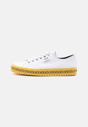 Weißer Canvas-Sneaker in niedriger Ausführung mit weißen Schnürsenkeln und einer gelben Sohle, die wie ein Maßband mit Größenmarkierungen gestaltet ist.