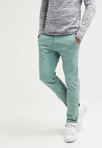 Mátově zelené, slim-fit kalhoty vyrobené z hladkého materiálu, v kombinaci se strukturovanou šedou košilí s dlouhým rukávem a bílými teniskami.
