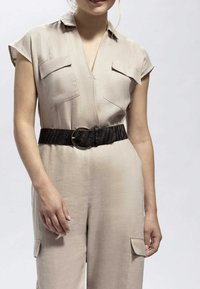 Beige jumpsuit med korta ärmar, två fickor med lappar, en V-ringning och ett strukturerat svart bälte med en cirkulär spänne.