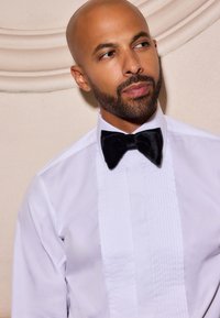 Camicia bianca da uomo con davanti plissettato, abbinata a un papillon in velluto nero. Il colletto è rigido e tradizionale nello stile.