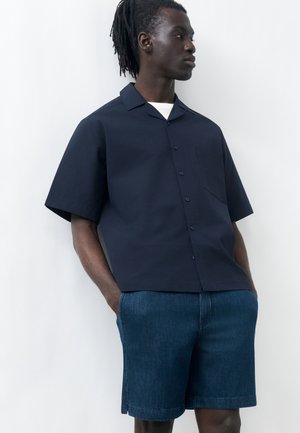 Man with dreadlocks wearing a navy short-sleeve button-up shirt and denim shorts, standing with hands in pockets against a white wall.