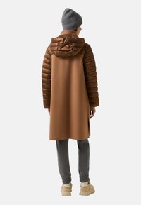 Dlouhá hnědá péřová bunda s kapucí, s kontrastním hladkým materiálem na zádech a rozparky po stranách, kombinováno se šedými kalhotami a béžovými botami.