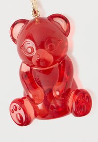 Punainen karhun muotoinen avaimenperä, joka on valmistettu läpinäkyvästä muovista. Siinä on pyöreät korvat, yksityiskohtaiset tassut ja kiiltävä pinta. Kiinnitetty kultainen metallirengas.