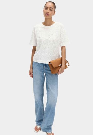 Kvinde iført hvid blomstermønstret kortærmet top, lyseblå brede jeans, hvide sandaler og holder en brun læder clutch.