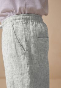 Un pantalon en lin gris clair avec un tissu texturé, une coupe décontractée, une taille élastique et une poche latérale unique sur le côté droit.