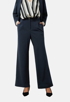 Pantaloni a gamba larga blu navy con una texture liscia, tasche frontali e vestibilità sartoriale. Abbinati a una blusa a righe e tacchi neri.