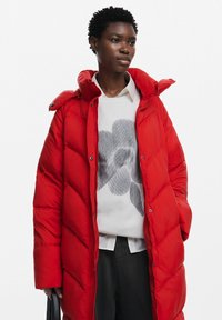 Abrigo acolchado rojo con un cuello alto, patrón acolchado y botones a presión. Llevado sobre un suéter ligero con un diseño floral gris.