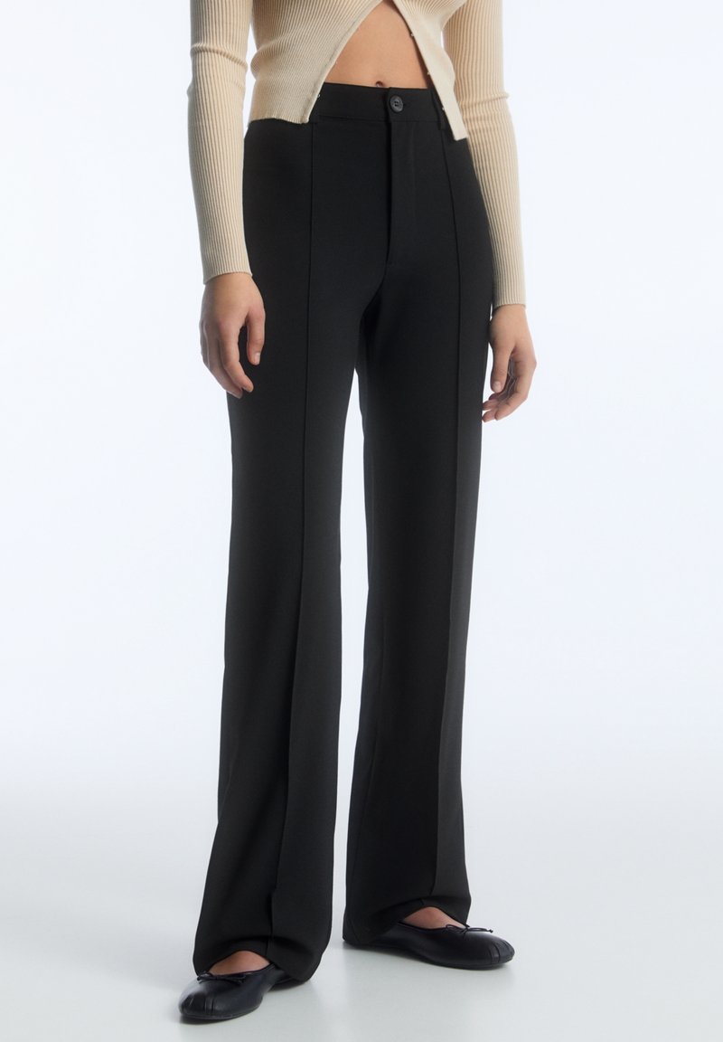 Personne portant un pantalon large noir taille haute, un cardigan court beige côtelé et des ballerines noires sur fond blanc.