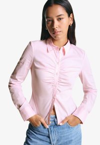 Camicia button-up rosa chiaro con colletto, caratterizzata da sottili strisce verticali e dettagli in tessuto arricciato lungo il davanti.