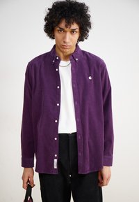 Paarse corduroy shirt met button-down kraag, voorzien van witte knopen en een logo op de borst. Gedragen over een wit t-shirt met zwarte broek.