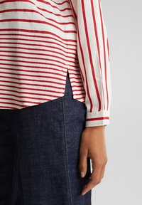 Blouse en coton rayée rouge et blanc avec manches longues, fentes latérales et poignets boutonnés, associée à un pantalon en jean foncé.
