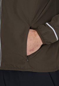 Veste légère marron avec une poche zippée, des poignets élastiques et des détails blancs le long des coutures. Texture lisse, design décontracté.