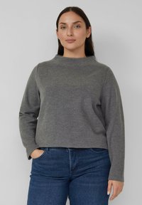 Grauer Pullover mit langen Ärmeln, hohem Kragen und lockerem Sitz, hergestellt aus einem weichen Strickstoff. Kombiniert mit dunkelblauen Jeans.