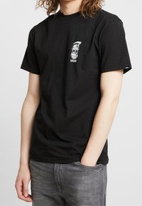 Musta puuvillainen t-paita, jossa valkoinen kallokuvio vasemmassa rinnassa ja "VANS"-teksti. Pyöreä pääntie, lyhyet hihat, standardi mitoitus.