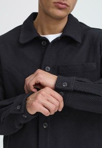 Black, corduroy shirt with a collar, buttoned placket, and pocket. Features textured fabric and contrasting black buttons.