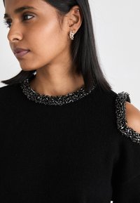 Μαύρος πλεκτός σκούφος με υφή από μαύρες χάντρες γύρω από το λαιμό και στον ώμο, με ανοιχτό σχέδιο στον ώμο.