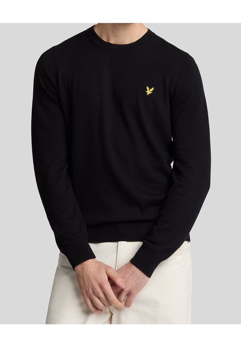 Schwarzer Strickpullover mit Rundhalsausschnitt, gerippten Bündchen und Saum; mit einem kleinen gelben Vogel-Emblem auf der linken Brust.