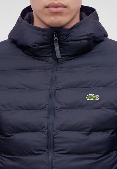 Homme portant une veste matelassée noire zippée avec une capuche et un petit logo crocodile vert sur la poitrine.