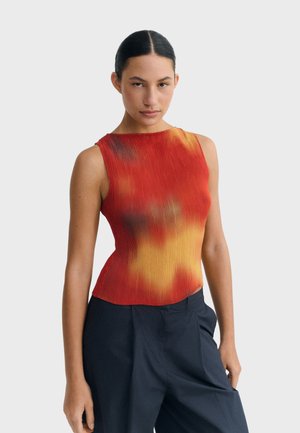 Kobieta z ciemnymi włosami związanymi do tyłu, ubrana w bezrękawnik w czerwono-pomarańczowym gradientowym kolorze oraz luźne ciemnoniebieskie spodnie, stojąca na jednolitym tle.