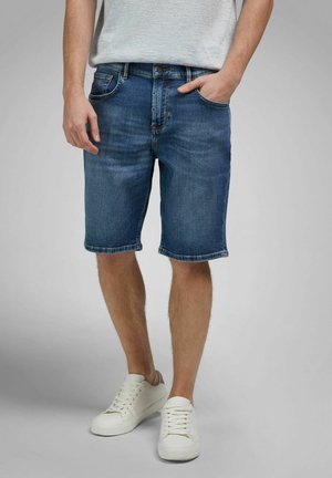 Mann trägt blaue Jeansshorts und weiße Turnschuhe, steht mit einer Hand in der Tasche vor einem einfarbigen Hintergrund.