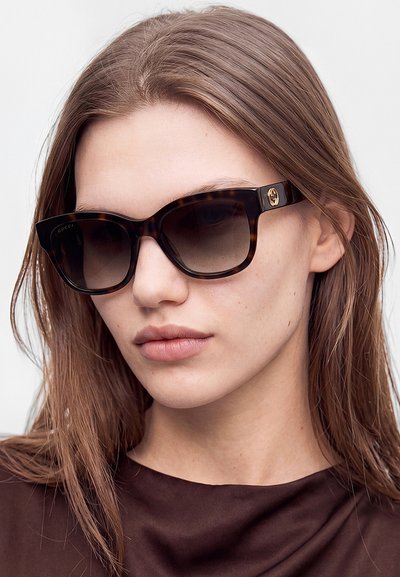 Donna con capelli lisci castano chiaro che indossa occhiali da sole Gucci con montatura tartarugata scura e una maglietta marrone su uno sfondo chiaro e uniforme.