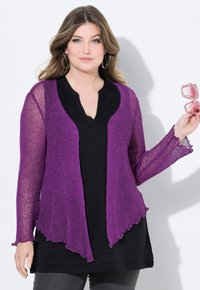 Lila stickad kofta med öppen fram, texturerad finish och vågiga kanter, lagd över en svart topp, i kombination med rosa solglasögon.