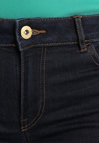 Donkerblauwe spijkerbroek met metalen knoopsluiting en oranje stiksels langs de naden, met een ontwerp van klassieke vijf zakken.