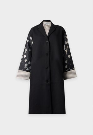 Femme debout portant un long manteau noir avec des manches à motif floral, un haut fleuri, un pantalon crème et des chaussures crème pointues sur un fond uni.