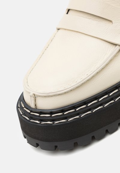 Kreemist nahast loafers, millel on paks must kummist tall. Omab õmmeldud ääre ja pilu detaili peal, rõhutades selle tekstureeritud viimistlust.
