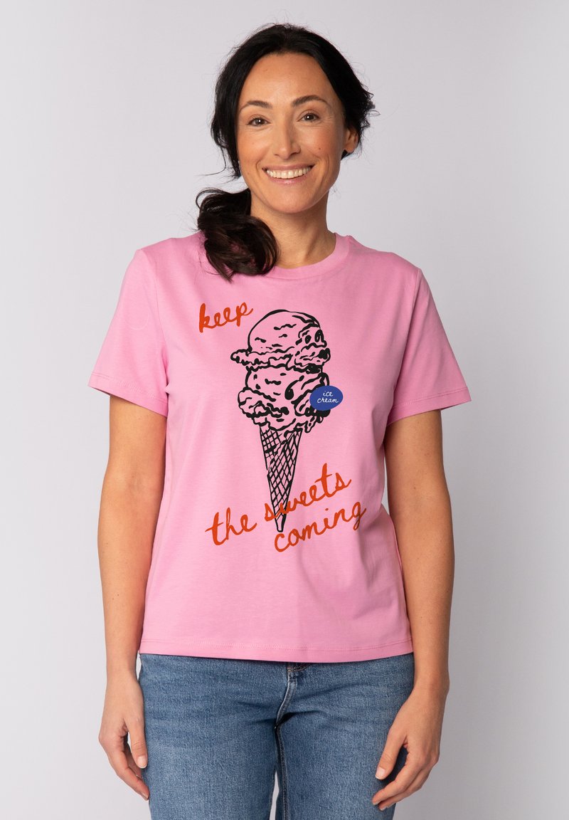 Pinker Baumwoll-T-Shirt mit einem Grafikdesign eines Eisbechers in schwarzer Umrandung und rot-orangem Text "Halt die Süßigkeiten nach." Kurze Ärmel, Rundhalsausschnitt.