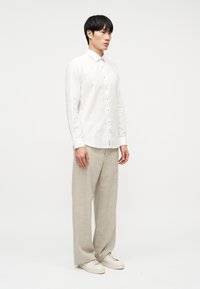 Άνδρας που φοράει λευκό πουκάμισο με κουμπιά, μπεζ παντελόνι με φαρδιά μπατζάκια και λευκά αθλητικά παπούτσια, στέκεται μπροστά σε απλό λευκό φόντο.