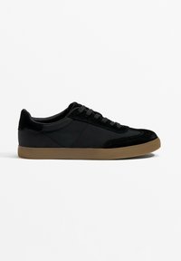 Black sneaker with suede and smooth synthetic upper, textured black laces, and tan rubber sole, featuring a low profile and rounded toe.