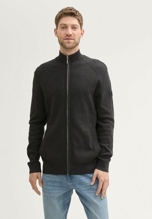 Tamno siva zip-up kardigan s rebrastom teksturom, visokim ovratnikom i bočnim džepovima. Nosite ga preko svijetloplavih traperica. Uređen kroj.