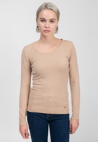Beige Langarmpullover mit rundem Ausschnitt, gerippten Bündchen und Saum. Weiche Textur, taillierter Schnitt. Goldenes Logo-Highlight an der Hüfte.