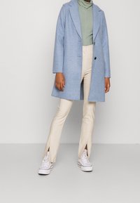 Cappotto in lana azzurro chiaro con rever a punta e un solo bottone, indossato su un dolcevita verde, abbinato a pantaloni beige con spacco e sneakers bianche.