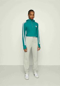 Sweatshirt de fecho em teal com riscas brancas nas laterais e um patch com o logótipo, combinado com calças de treino cinza claro e ténis brancos.