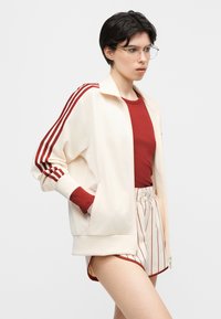 Jeune personne portant une veste crème avec des rayures rouges, un t-shirt rouge côtelé, et un short crème avec des rayures verticales rouges, debout sur un fond uni.