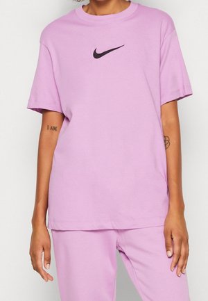 Person iført en lyse lilla Nike T-shirt og matchende bukser, der viser armtatoveringer med delvis tekst og et blomsterdesign.