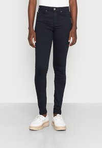 Jeans skinny de color azul oscuro con un acabado suave, que presentan un diseño clásico de cinco bolsillos y un cierre de botón plateado. Llevados con zapatillas blancas.
