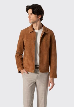 Muž v hnědém semišovém kabátu přes bílou košili a béžových kostkovaných kalhotách, stojící s jednou rukou v kapse, hledící doleva.