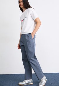 Wit T-shirt met rood en blauw logo, grijze op maat gemaakte broek en zilveren sportschoenen. Glad materiaal en relaxte pasvorm.